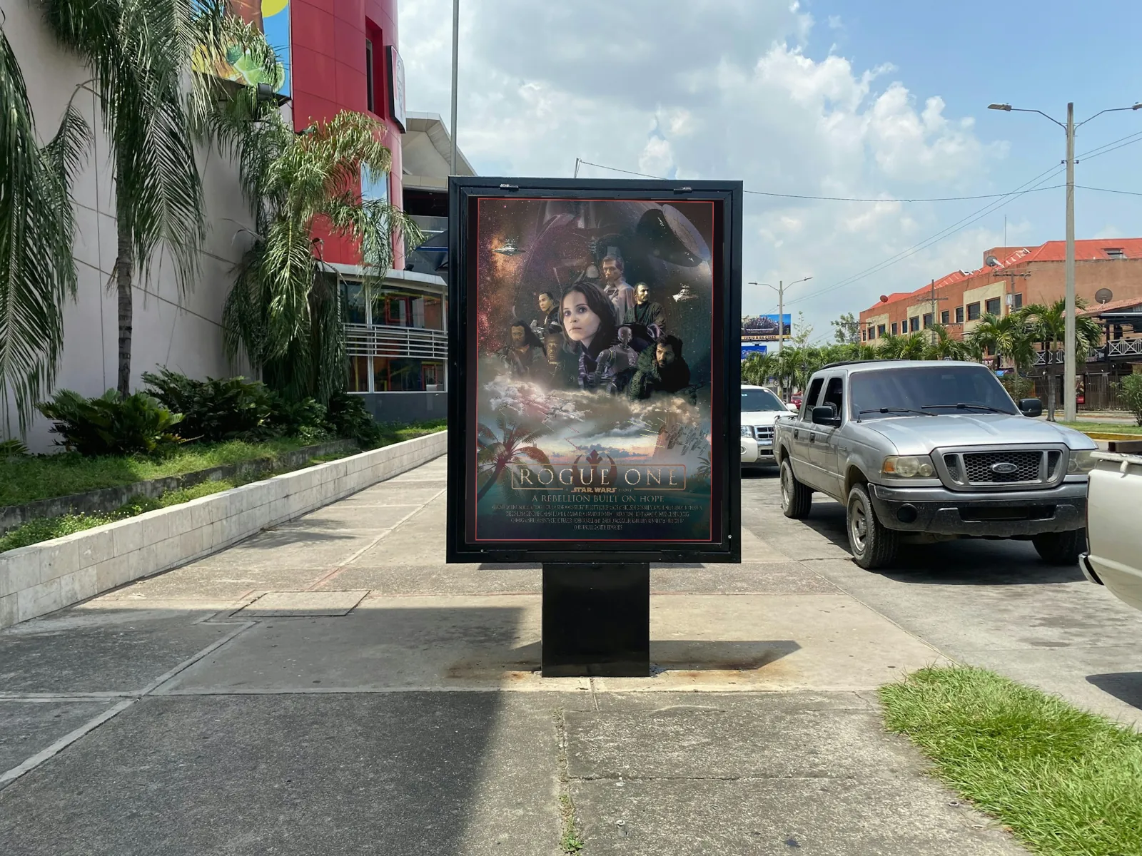 El cartel de «Rogue One» en un mupi a pie de calle, junto a coches aparcados y palmeras.